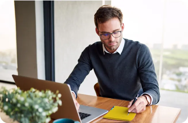 Man in front of a computer writing on a notebook working on a website.